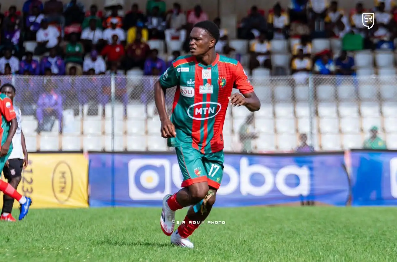 Soccer player in teal and red kit number 17 runs with the ball on a grass field during a match, stands in the background.