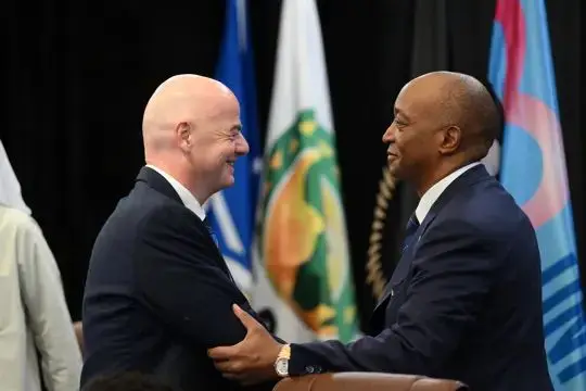 Two bald men in suits shake hands and smile at a formal event, with flags in the background. Two bald men in suits shake hands and smile at a formal event, with flags in the background.
