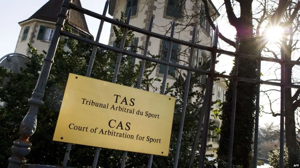 Gold sign on a black iron fence reading 'TAS Tribunal Arbitral du Sport' and 'CAS Court of Arbitration for Sport' with a building in the background and sunlight glare.