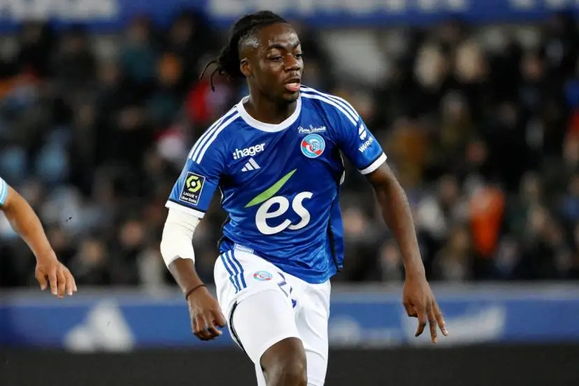 A soccer player in a blue jersey sprinting on the field during a match, arms ready, crowd blurred in the background.