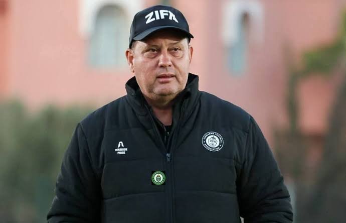 Middle-aged man in a black zip-up jacket with badges and a ZIFA cap, standing outdoors and looking toward the camera.