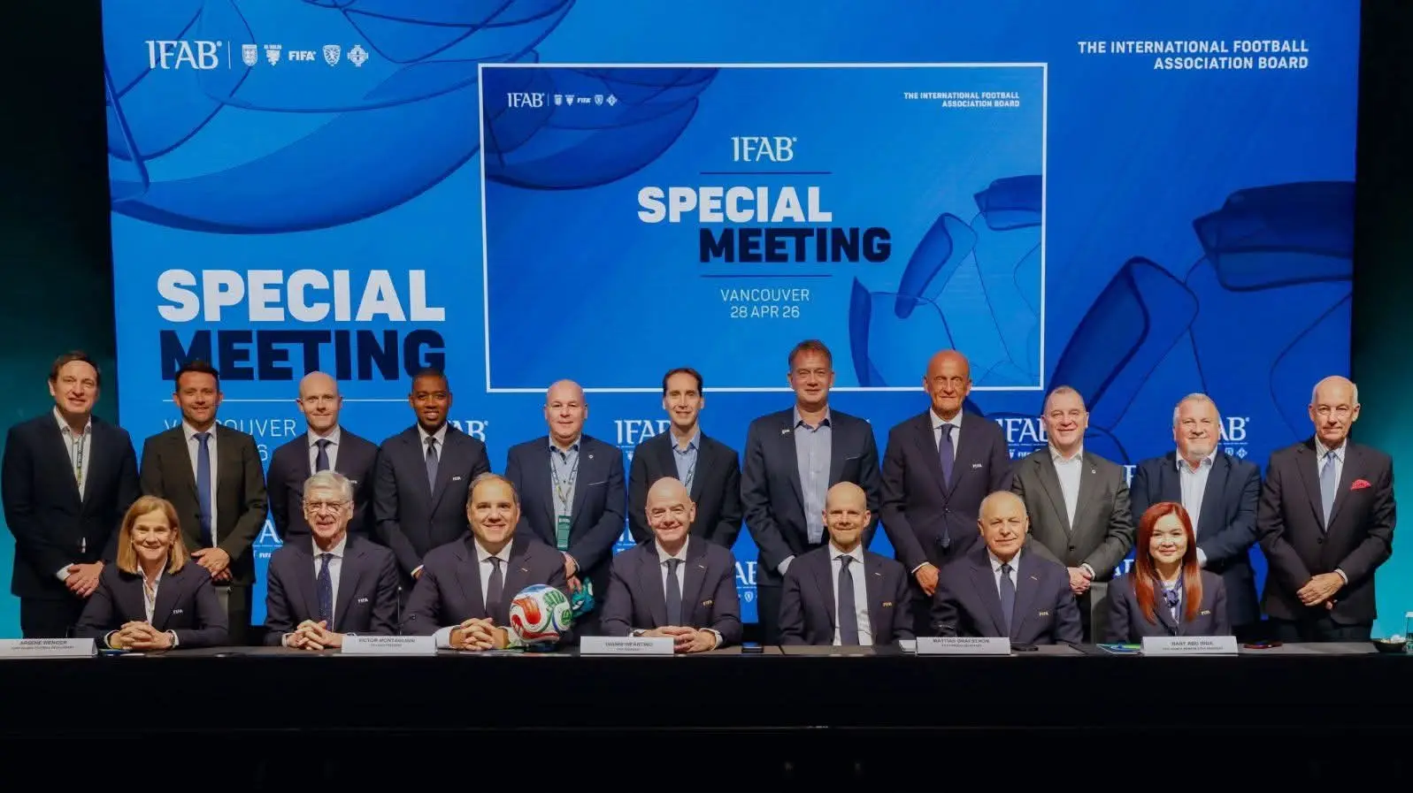 Group of formally dressed officials posing for a photo at a press conference in front of a blue backdrop reading 'SPECIAL MEETING' with a football on the table center-right and nameplates in front of seated individuals. Group of formally dressed officials posing for a photo at a press conference in front of a blue backdrop reading 'SPECIAL MEETING' with a football on the table center-right and nameplates in front of seated individuals.