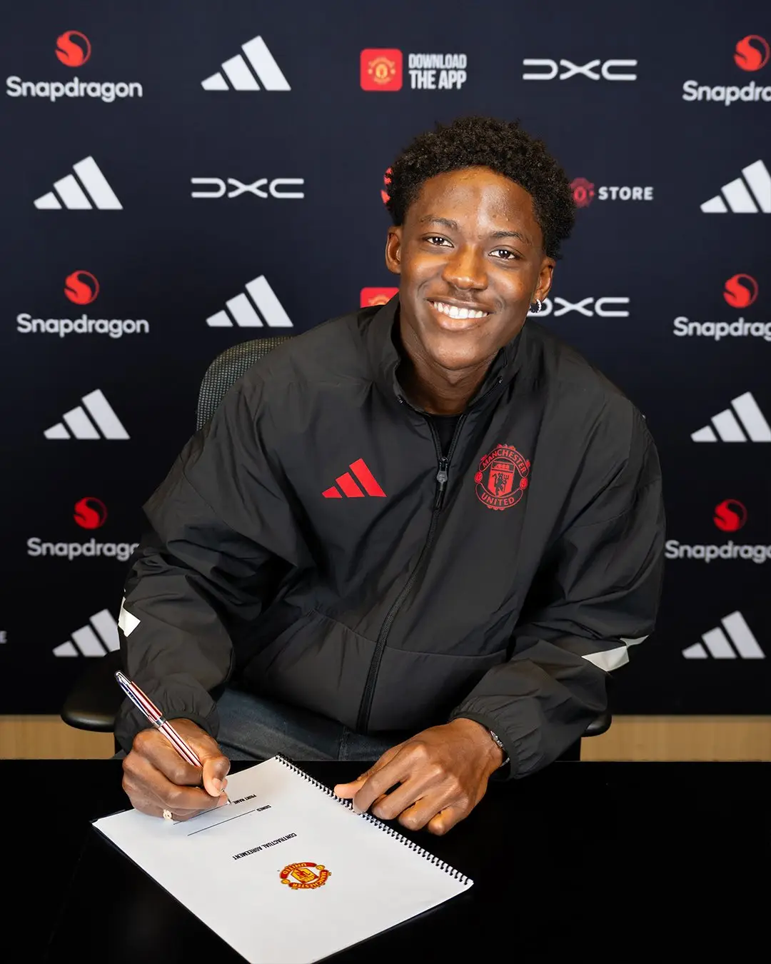 Smiling man in a Manchester United jacket signs a contract at a desk during a signing event, backdrop with sponsor logos.