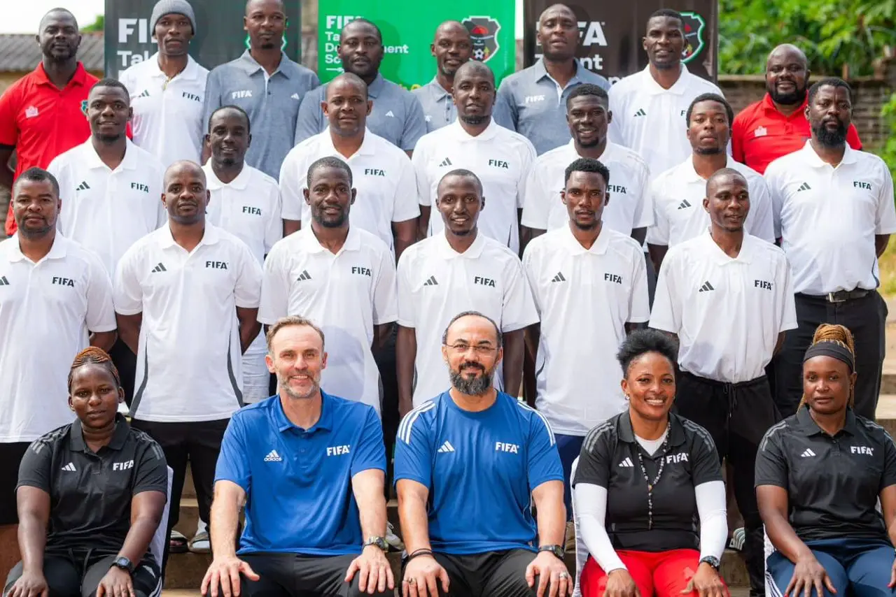 Group portrait of FIFA staff and coaches on outdoor steps; front row seated in blue FIFA shirts, others in white FIFA polo shirts, with a few women in darker tops in the front row. Group portrait of FIFA staff and coaches on outdoor steps; front row seated in blue FIFA shirts, others in white FIFA polo shirts, with a few women in darker tops in the front row.