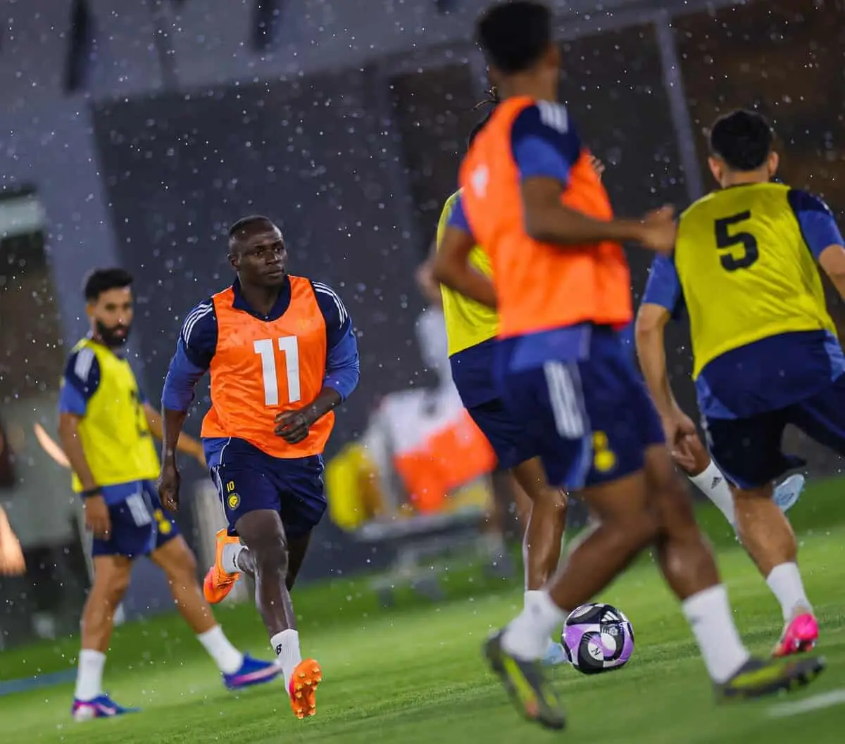 Soccer players train on a wet field at night, wearing orange and yellow bibs as a ball moves between them in the rain
