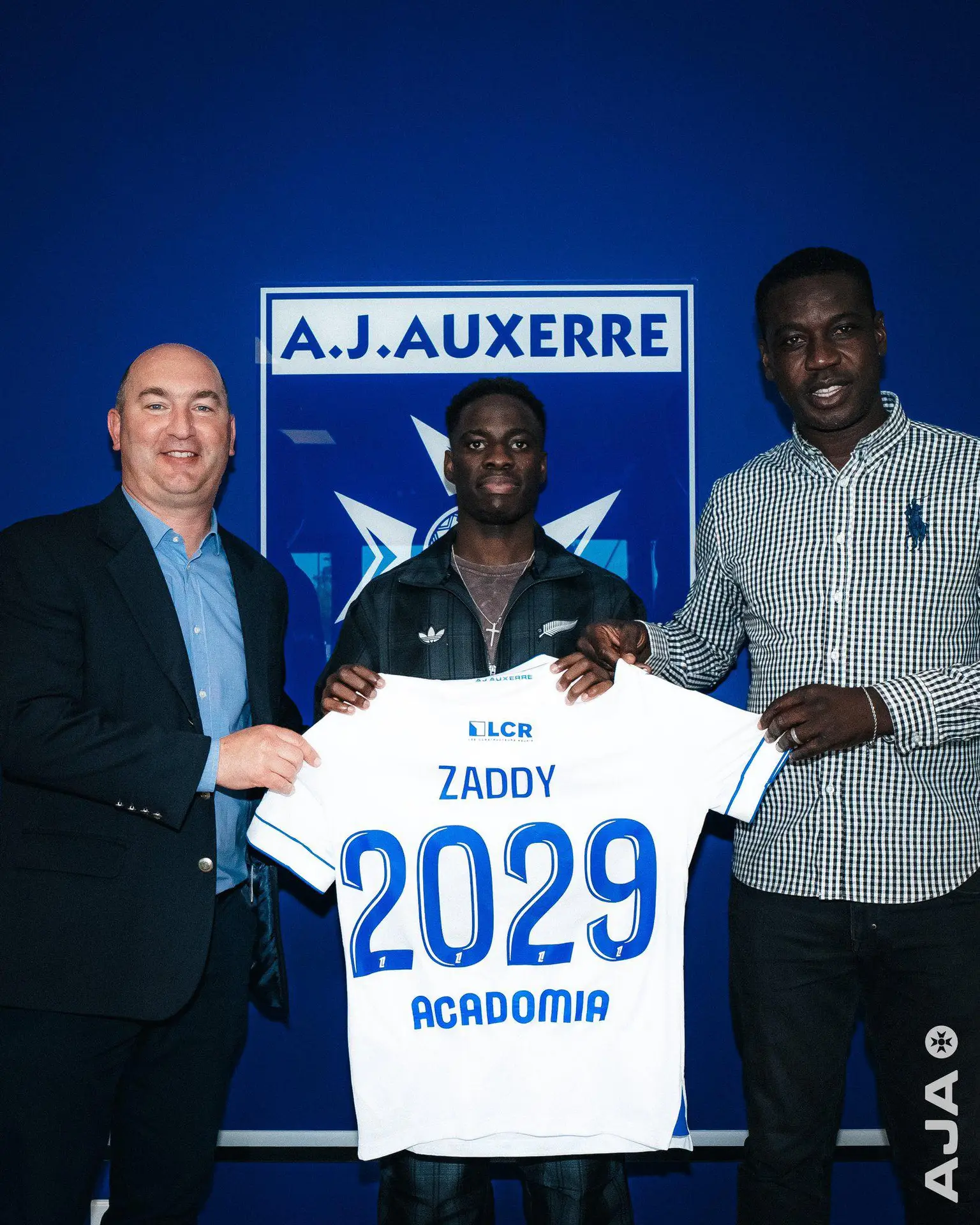 Three men pose in front of a blue A.J. Auxerre backdrop, holding a white jersey between them.