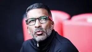 Man with short hair and a trimmed beard wearing glasses and a black jacket, with red stadium seats blurred in the background.