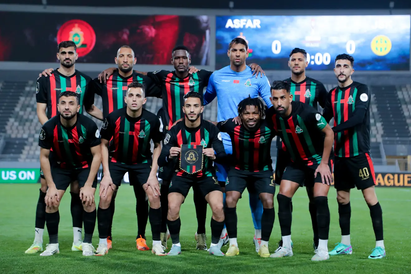 Soccer team in black jerseys with red and green vertical stripes posing for a group photo on the field, with a blue stadium backdrop behind them.