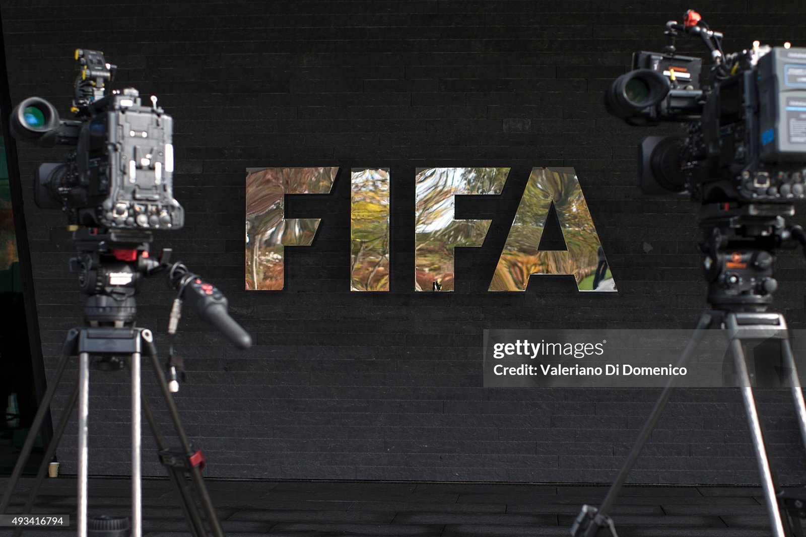 Two professional broadcast cameras on tripods facing a dark brick wall with large mirrored letters in the background.