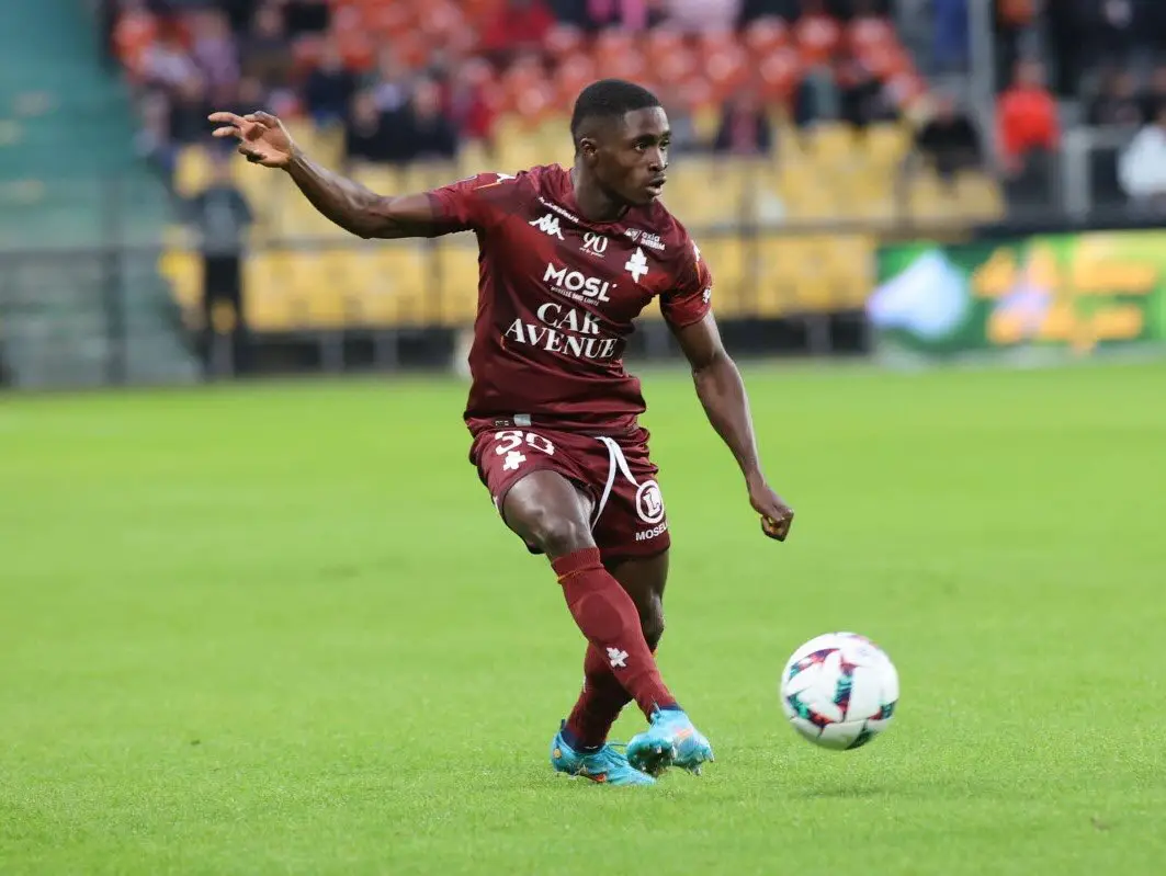Soccer player in a maroon kit kicks a ball on a green field during a match, stadium and fans in the background. Soccer player in a maroon kit kicks a ball on a green field during a match, stadium and fans in the background.