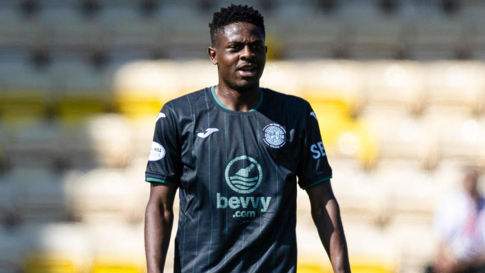 Soccer player in a dark teal kit stands on the field during a match, with a team crest and sponsor logo on the chest. Soccer player in a dark teal kit stands on the field during a match, with a team crest and sponsor logo on the chest.