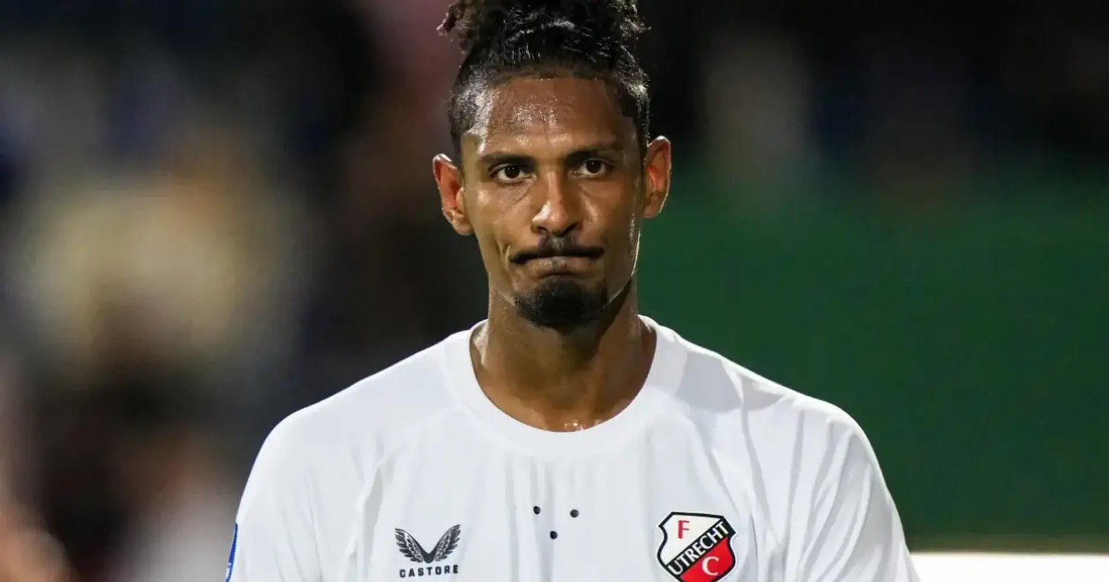 Close-up of a male footballer in a white FC Utrecht jersey, looking ahead with a neutral expression, stadium lights in the background Close-up of a male footballer in a white FC Utrecht jersey, looking ahead with a neutral expression, stadium lights in the background