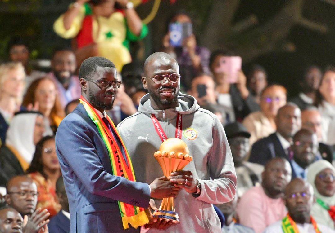 Two men on stage exchange a trophy at an awards ceremony, with a diverse audience clapping in the background.