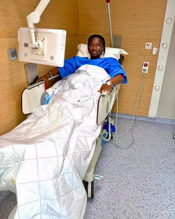 Patient in a hospital bed, blue gown, white quilt up to the chest, in a room with medical equipment overhead.
