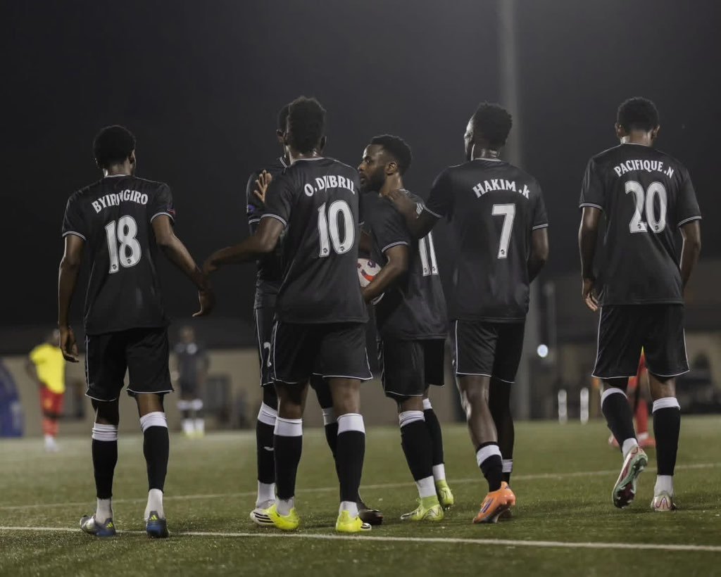 Group of five soccer players in dark uniforms walk together on a lit field, backs to the camera at night.
