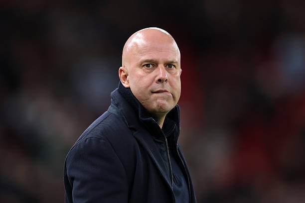 Sidelined, bald man in a dark coat gazes seriously toward the camera on a sports field.