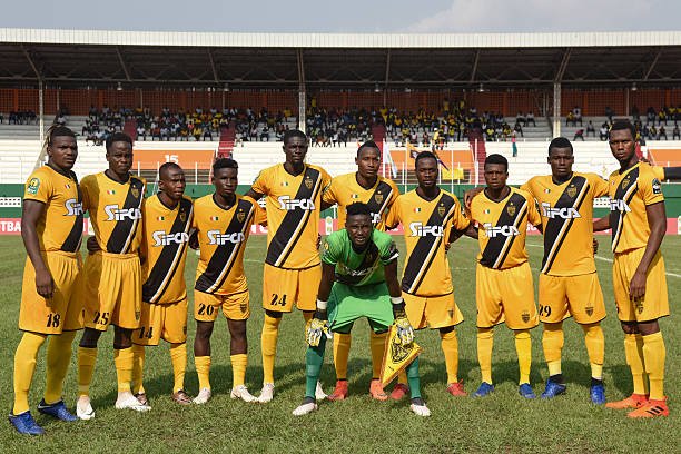 Team of eleven soccer players in yellow uniforms with black diagonal stripe, posing with goalie in green at center on a pitch.