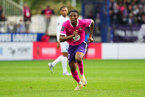 Female soccer player in a pink jersey and purple shorts sprinting on a grass football pitch during a match.