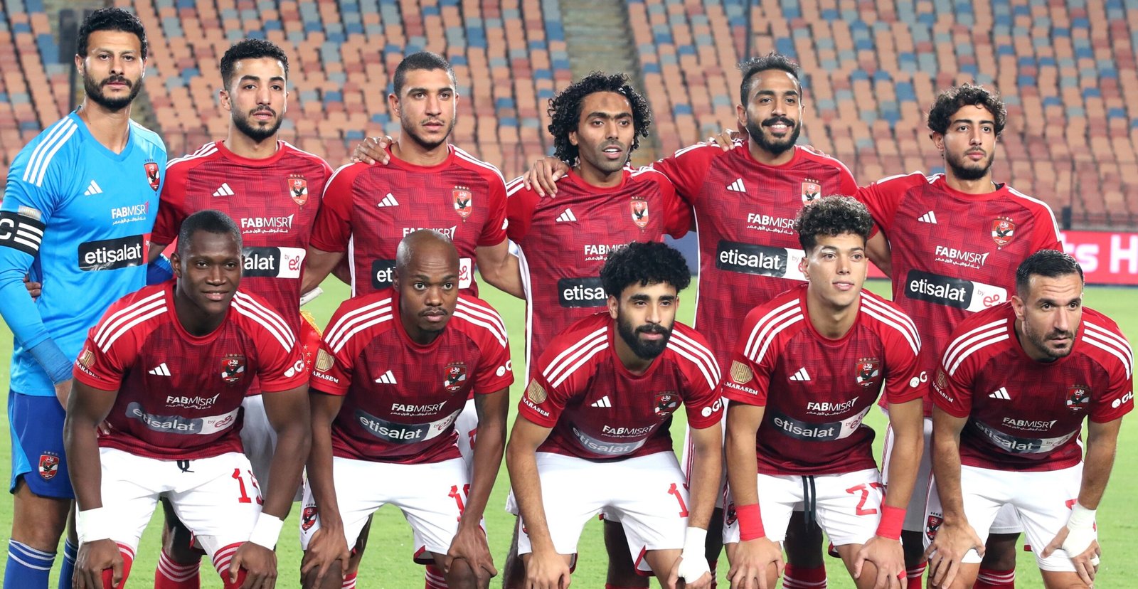 Group of soccer players in red jerseys posing for a team photo on the field, goalkeeper in blue at left, stadium seats in background.