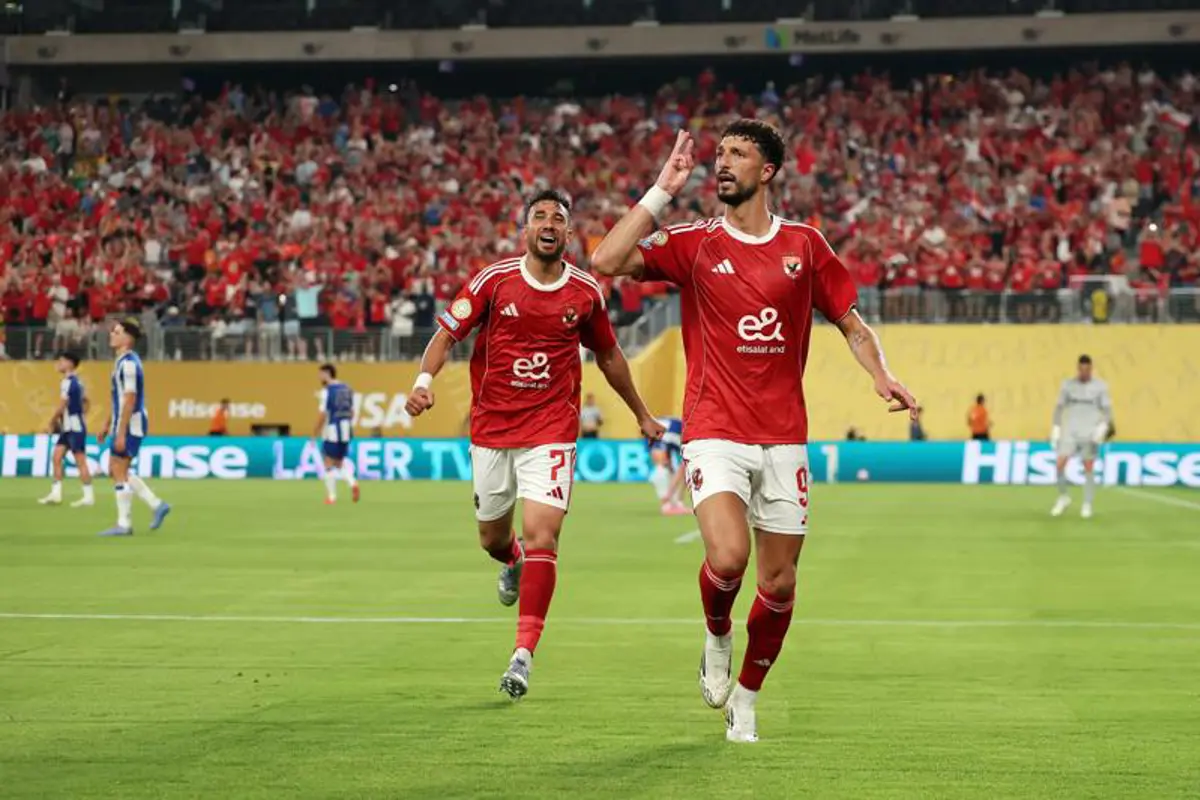 Two Egyptian football players in red jerseys celebrate a goal on a crowded stadium pitch with fans in red in the stands.