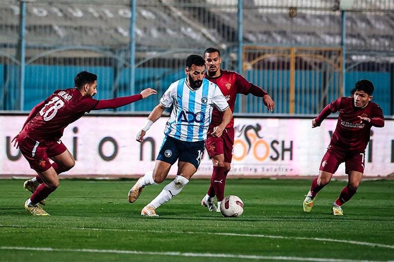 Soccer player in light-blue striped jersey dribbles the ball while three opponents in maroon pursue him on a green field stadium backdrop.
