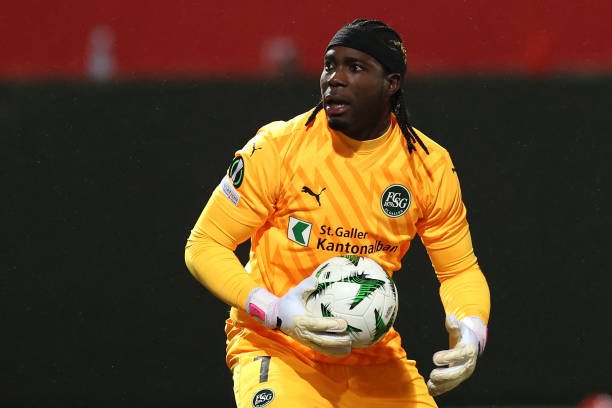 Soccer goalkeeper in an orange kit holding a ball with gloved hands during a match in a stadium.