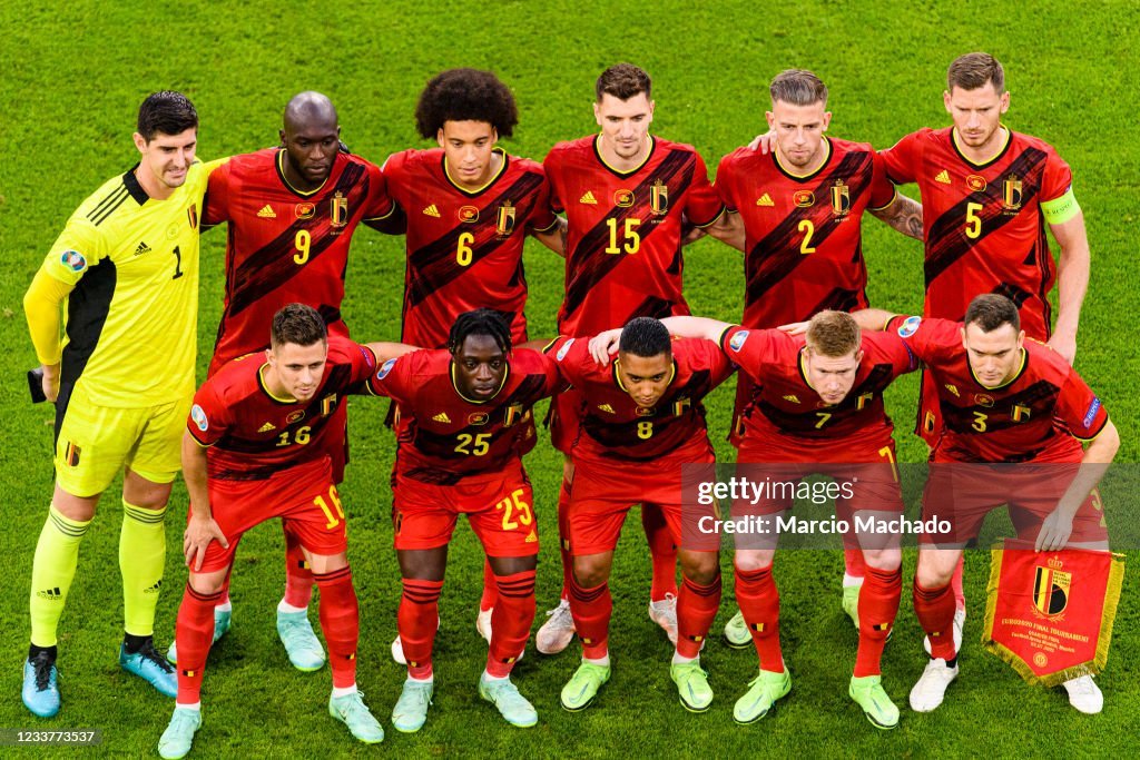 Belgian national football team teammates pose for a pre-match lineup on the green pitch, in red jerseys with yellow numbers and a yellow-clad goalkeeper on the left