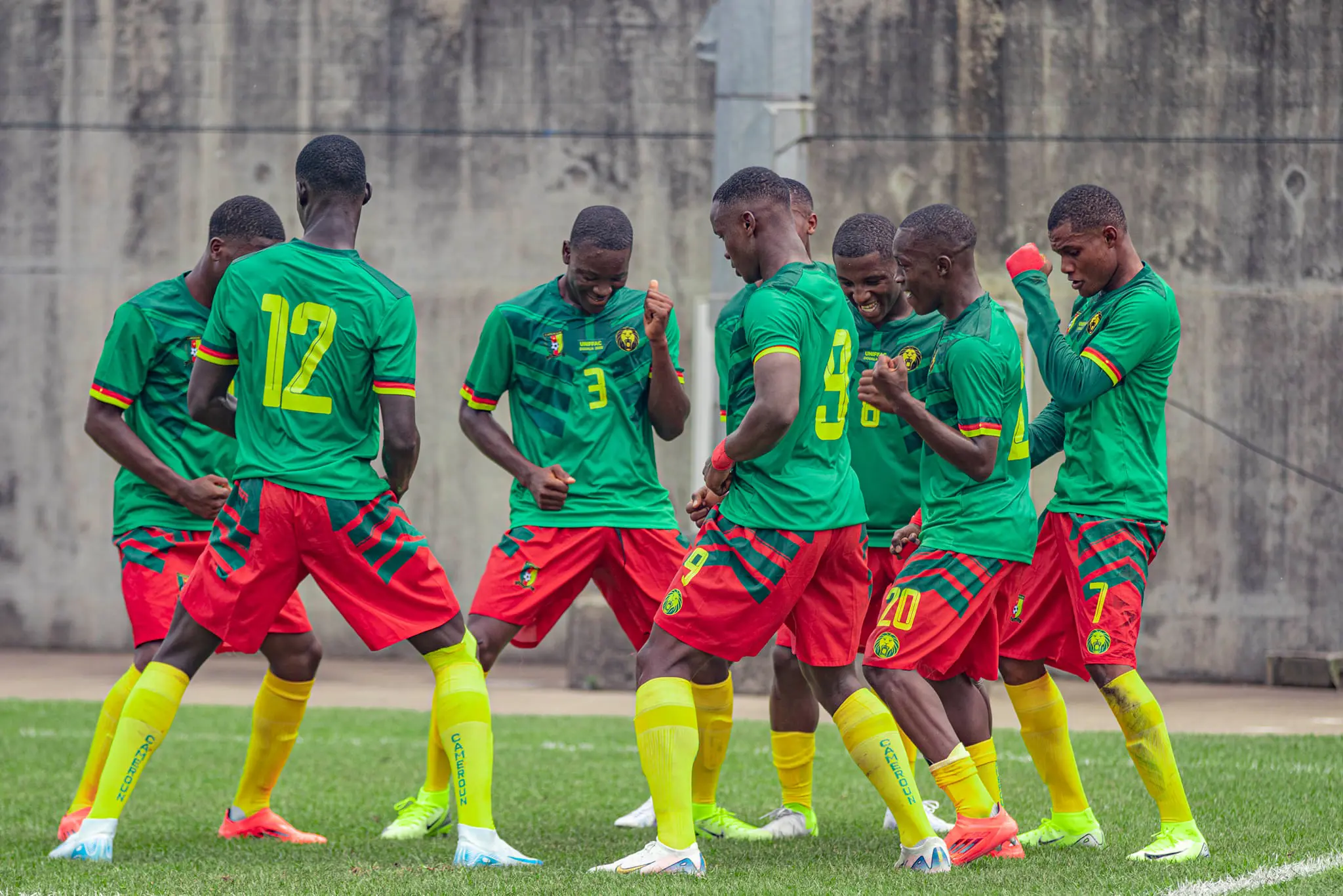 Soccer players in green uniforms with red shorts celebrate together on a grass field, forming a row with backs to the camera. Soccer players in green uniforms with red shorts celebrate together on a grass field, forming a row with backs to the camera.