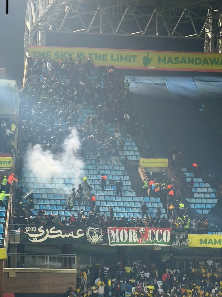 Crowd of soccer fans in blue seats at a stadium, with banners (Morocco, Arabic script) and a puff of smoke near the left side. Crowd of soccer fans in blue seats at a stadium, with banners (Morocco, Arabic script) and a puff of smoke near the left side.