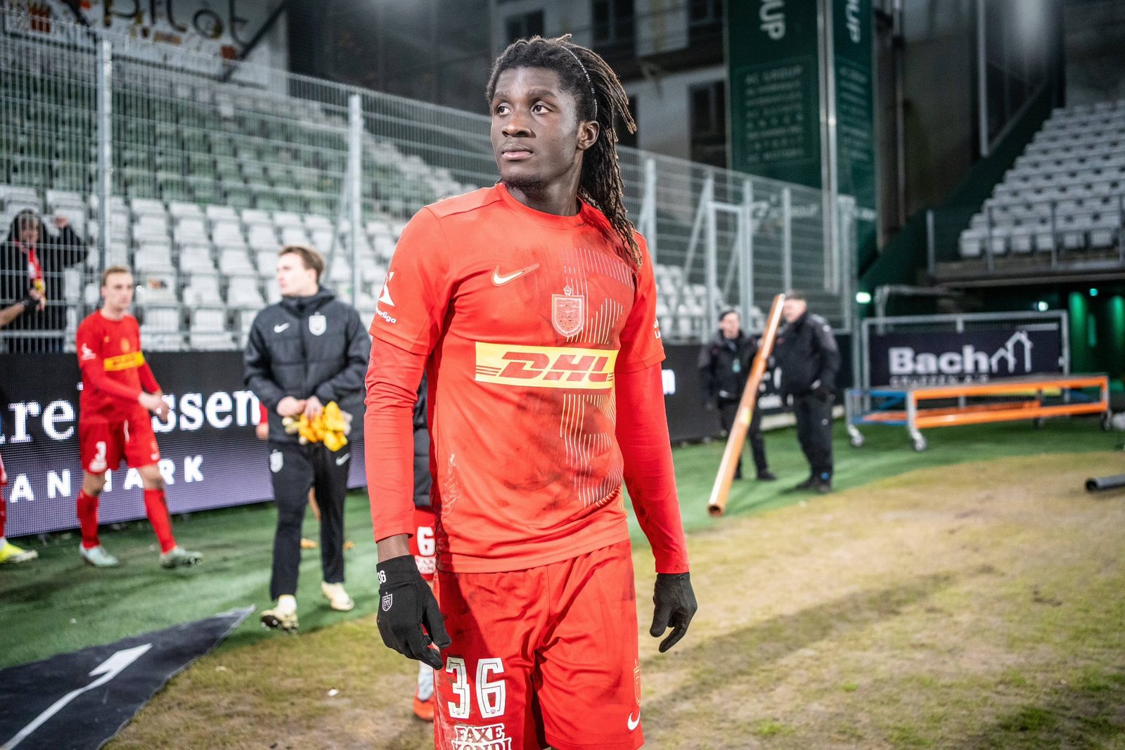 Soccer player in a red jersey and gloves stands on a field, looking off to the left with empty stadium seats behind him.