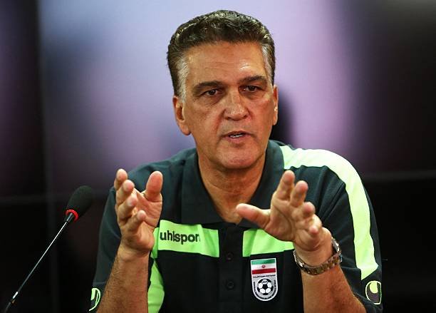Man in a sports polo with neon green stripes, speaking into a microphone and gesturing with his hands at a press event.