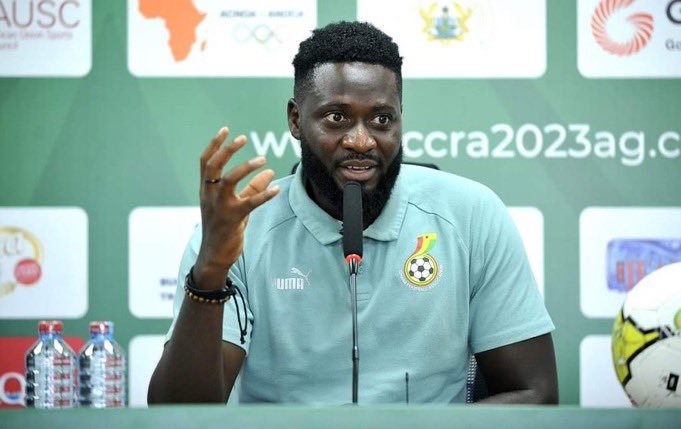 Man at a press conference wearing a light teal polo with a crest, speaking into a microphone and gesturing with his hand, with banners in the background and a soccer ball visible on the right.