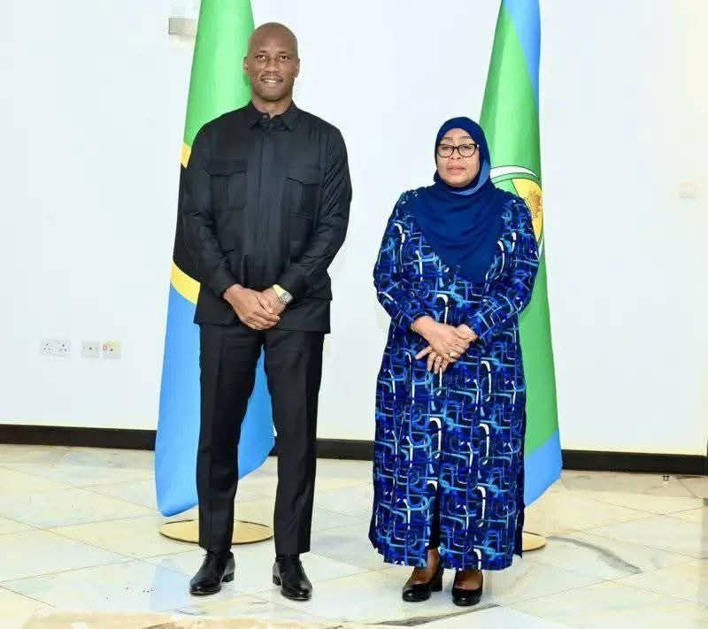 Man in black suit and woman in blue patterned dress with hijab standing indoors, with flag banners in the background. Man in black suit and woman in blue patterned dress with hijab standing indoors, with flag banners in the background.