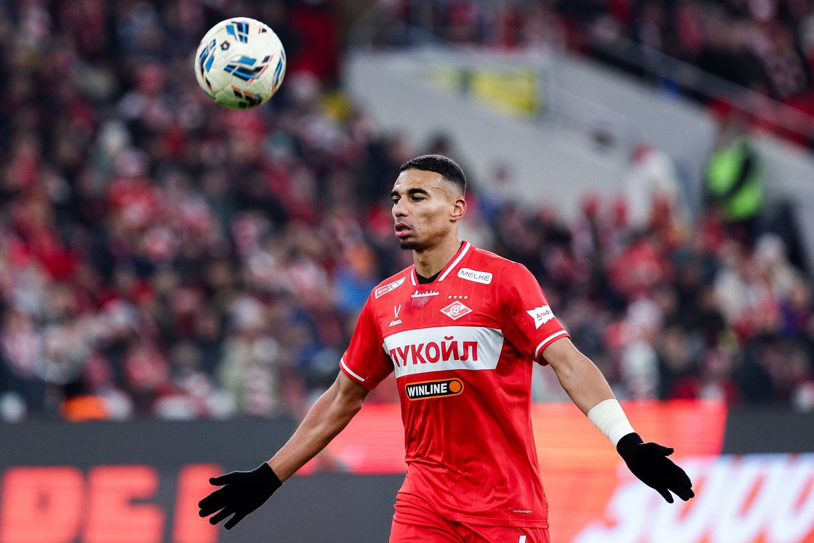 Soccer player in a red kit watches a ball drop from the air during a match with a blurred stadium crowd behind him.