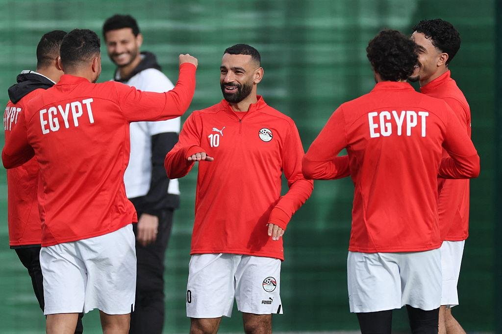 Egyptian football players in red training tops chat and stretch on a practice field.