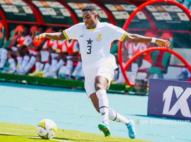 Soccer player in a white kit with number 3 kicking a ball during a match, with teammates on the bench in red seats behind. Soccer player in a white kit with number 3 kicking a ball during a match, with teammates on the bench in red seats behind.