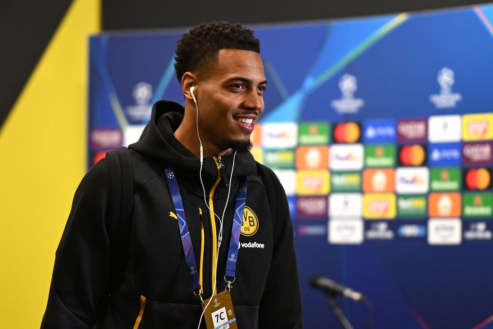 Smiling man wearing a black hoodie with Borussia Dortmund badge and a lanyard, listening with white earphones at a press conference backdrop.