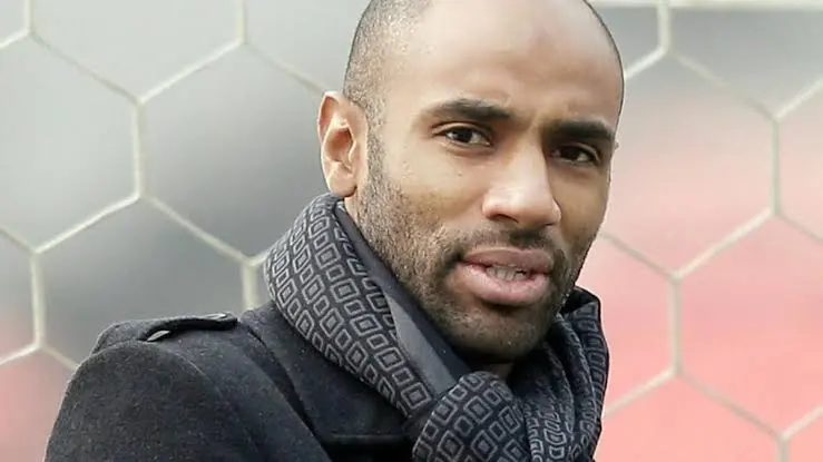 Close-up of a man with a short beard wearing a dark coat and a patterned scarf, outdoors with a hexagonal fence in the background. Close-up of a man with a short beard wearing a dark coat and a patterned scarf, outdoors with a hexagonal fence in the background.