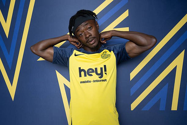 Athlete in a yellow and blue soccer jersey posing with hands behind head against a geometric blue and yellow backdrop, brand 'hey!' visible on chest