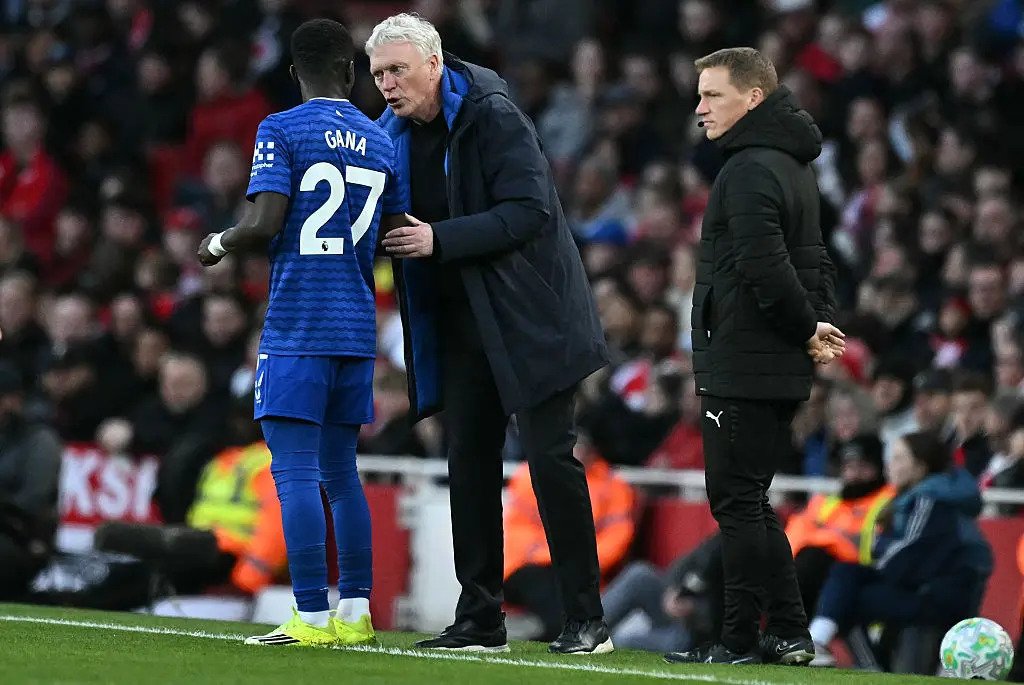 Football coach in a dark coat speaks to a player wearing blue kit and number 27 on the edge of the pitch, with another coach watching nearby.