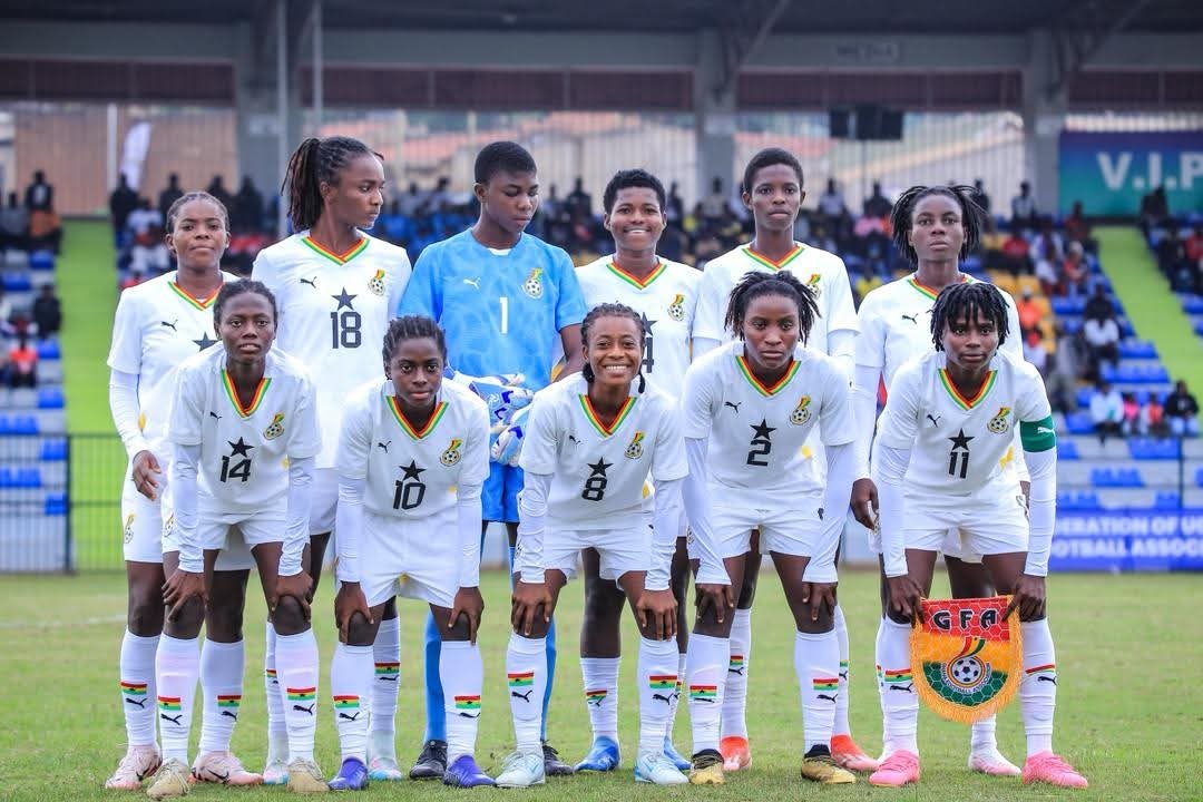 Girls' soccer team in white uniforms lined up for a group photo on the field, goalkeeper in blue at center back, one player holding a colorful banner. Girls' soccer team in white uniforms lined up for a group photo on the field, goalkeeper in blue at center back, one player holding a colorful banner.