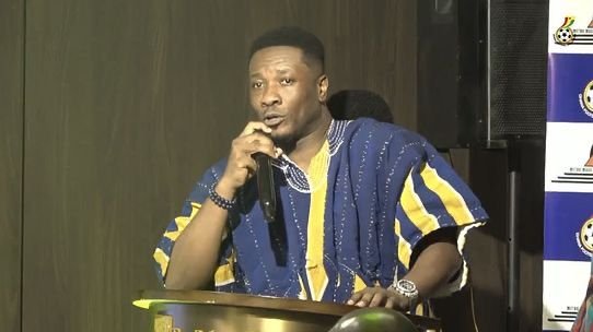 Man in blue and yellow patterned attire speaks into a handheld microphone at a lectern on a stage.