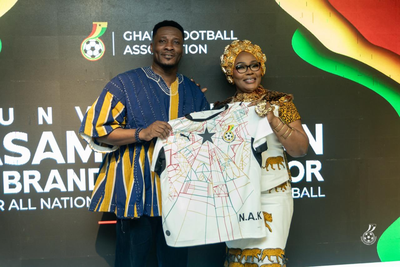 Two Ghanaian adults pose and display a white printed shirt with abstract colorful lines and a star, at a Ghana Football Association event backdrop with bold text behind them.