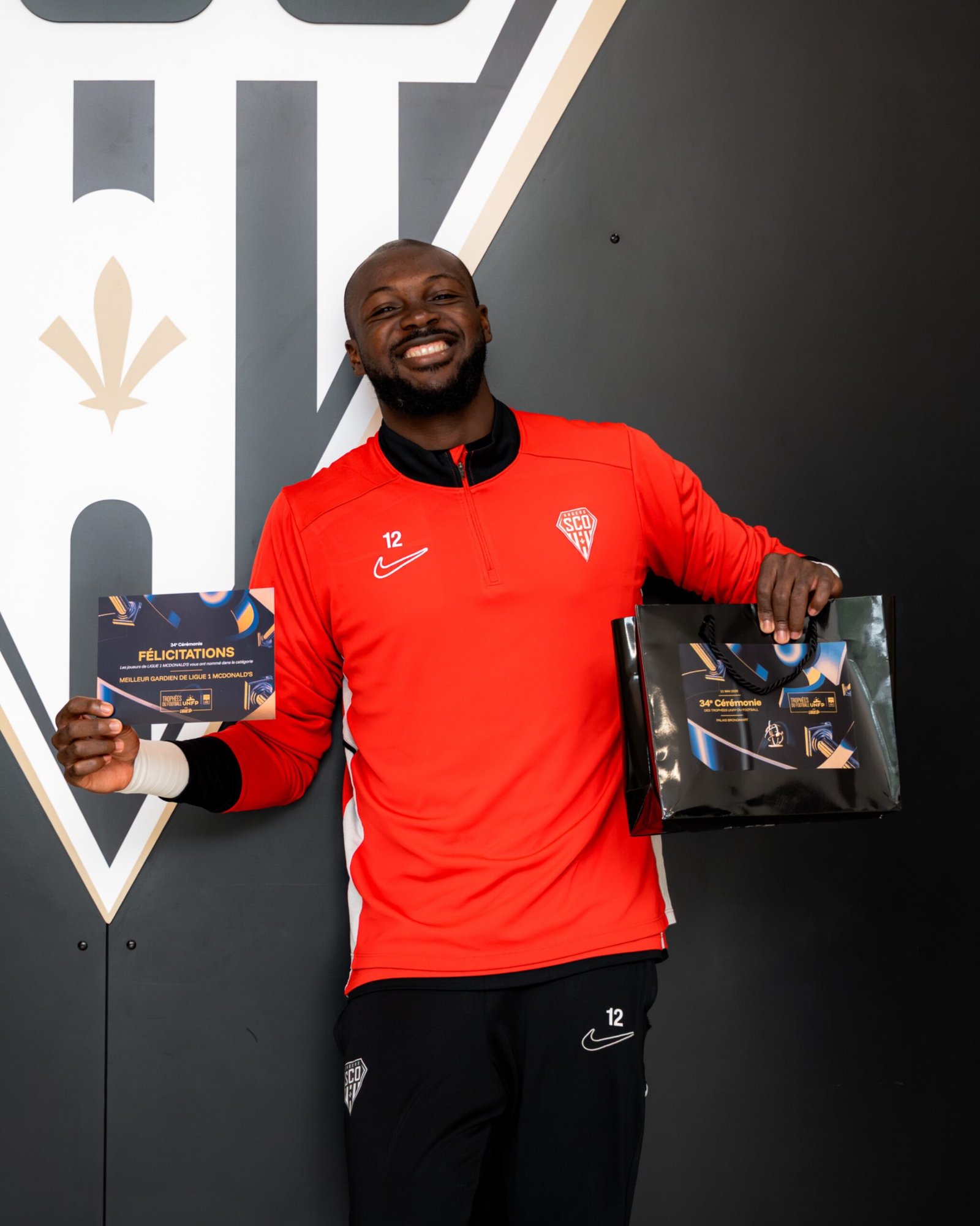 Smiling man in a red sports jersey holds an award certificate and a gift bag in front of a large logo backdrop.