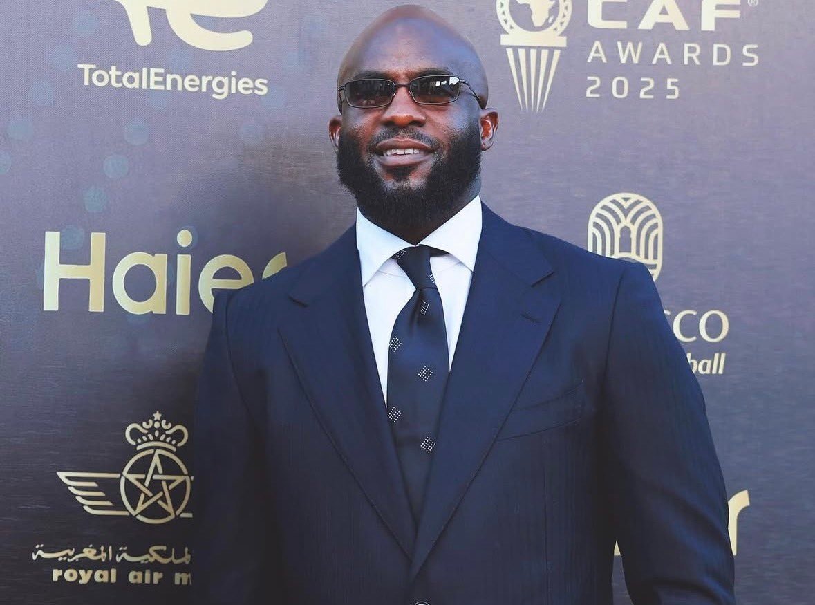 Man wearing a dark navy suit and sunglasses posing on a red carpet at an awards event, with sponsor logos on the backdrop behind him.
