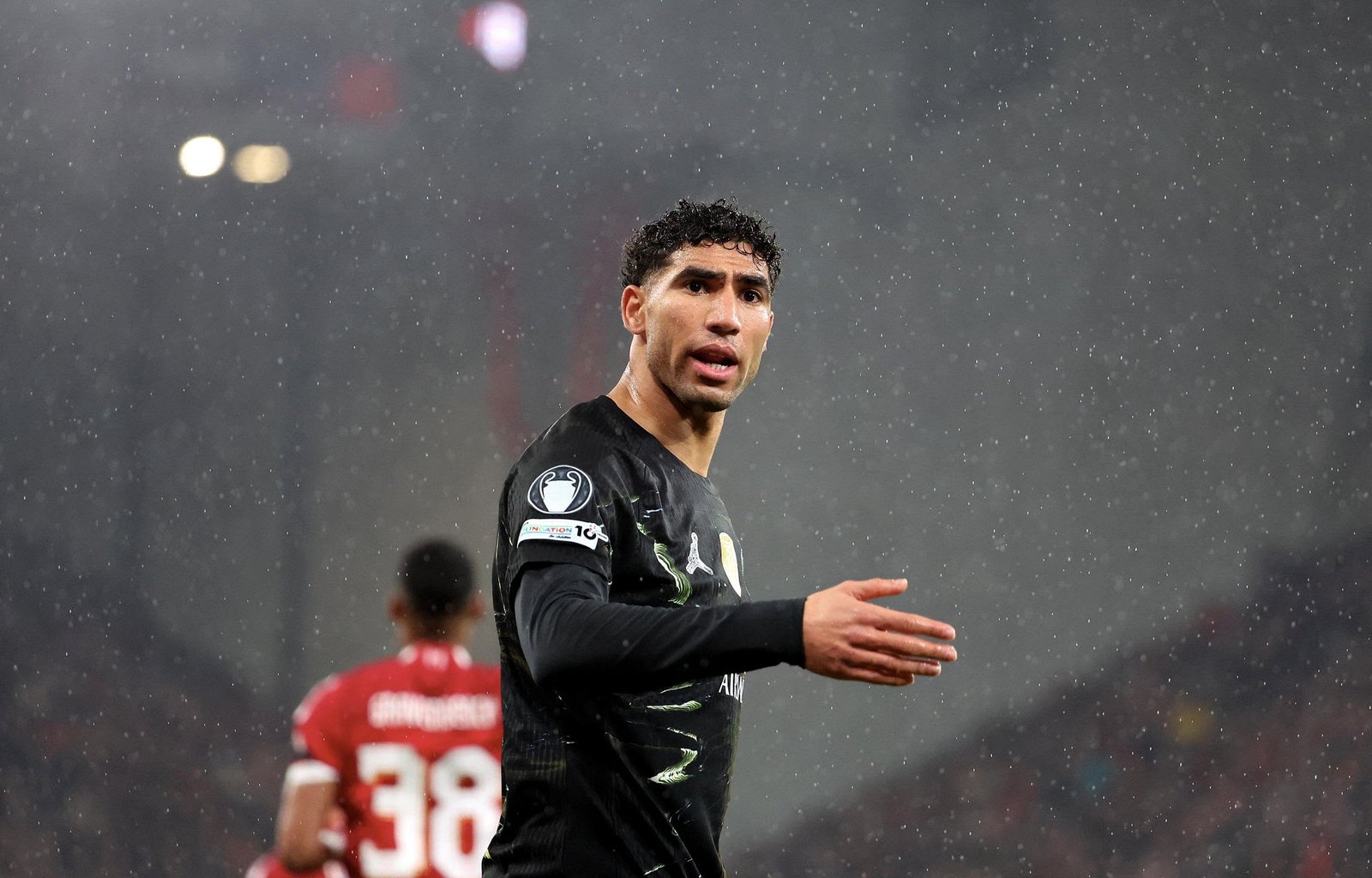 Footballer in a black kit gesturing in heavy rain on the pitch, with blurred players in red in the background.