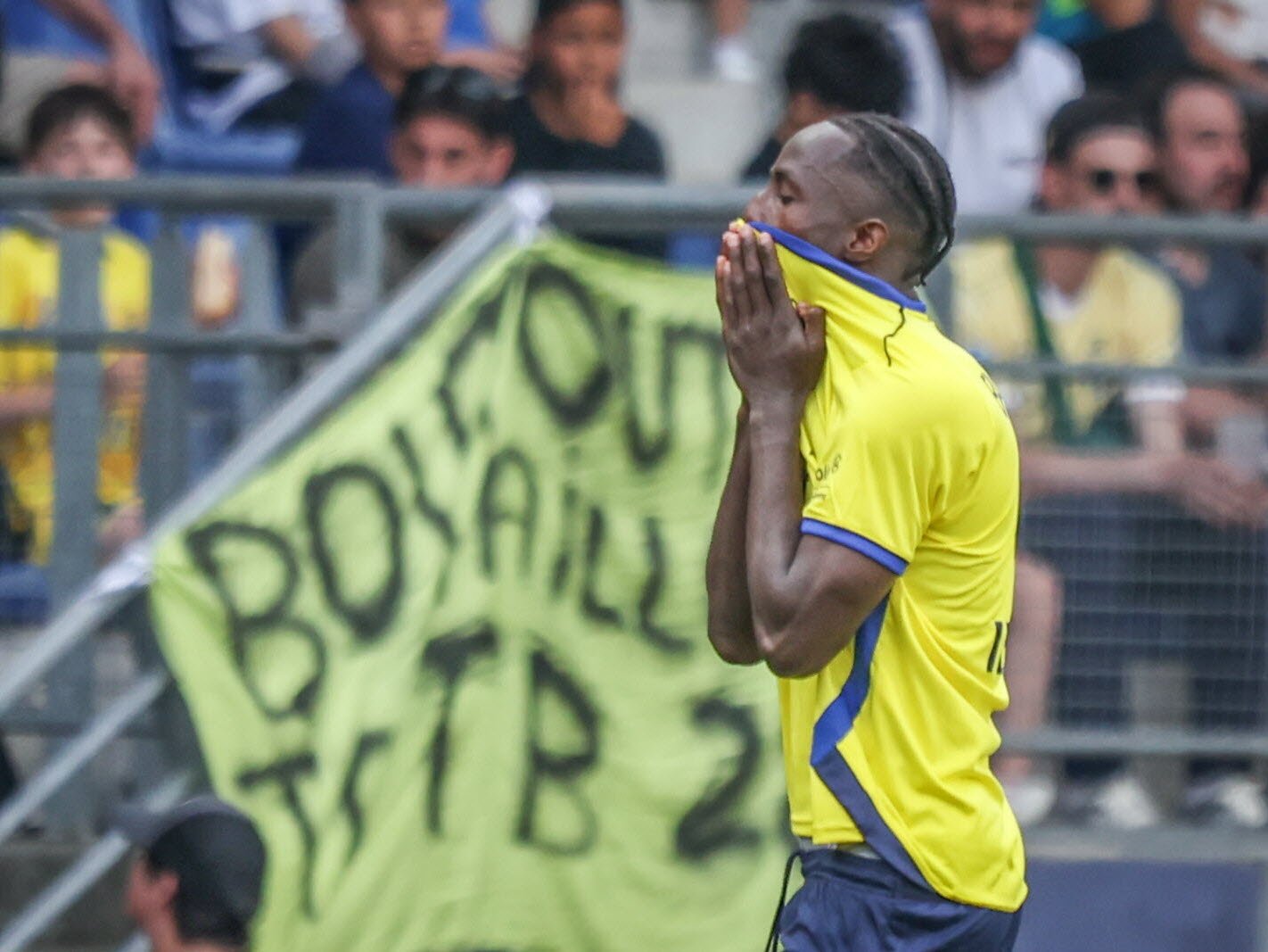 Soccer player in a yellow jersey covers his face with his hands on the pitch, with a bright green banner and spectators in the background.