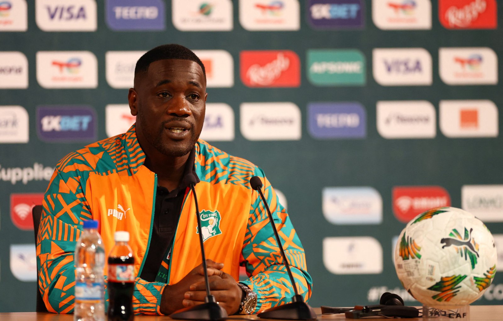 Man in a bright orange-teal patterned jacket speaks at a press conference with sponsor logos behind him and a soccer ball on the table Man in a bright orange-teal patterned jacket speaks at a press conference with sponsor logos behind him and a soccer ball on the table