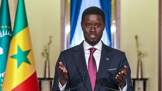 Man in a dark suit and magenta tie speaks at a podium with two microphones, Senegal flags behind him.