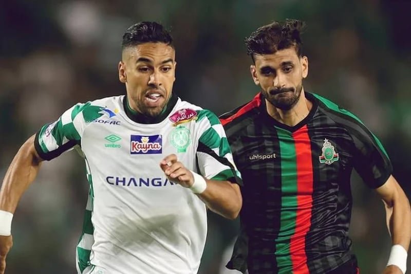 Two male soccer players sprint side by side during a match; one in a white and green kit, the other in a black kit with green and red stripes.